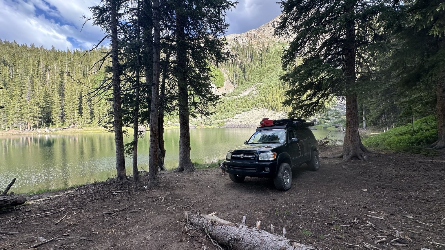 The black Sequoia parked beneath trees beside a mountain lake.